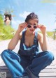 A woman sitting on a wooden dock wearing overalls and sunglasses.