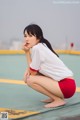 A woman in a white shirt and red shorts crouching on a tennis court.